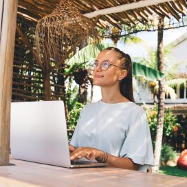 Young woman in casual t shirt and glasses sitting at wooden table in gazebo near palms working remotely on netbook